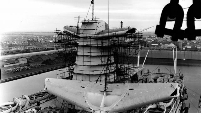 Dans le quai d'armement du bassin de Penhoët à Saint-Nazaire, des ouvriers des Chantiers de l’Atlantique procèdent à l'embarquement d'un aileron de cheminée du paquebot France (1962).