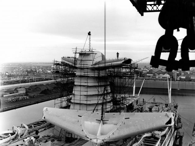 Dans le quai d'armement du bassin de Penhoët à Saint-Nazaire, des ouvriers des Chantiers de l’Atlantique procèdent à l'embarquement d'un aileron de cheminée du paquebot France (1962).